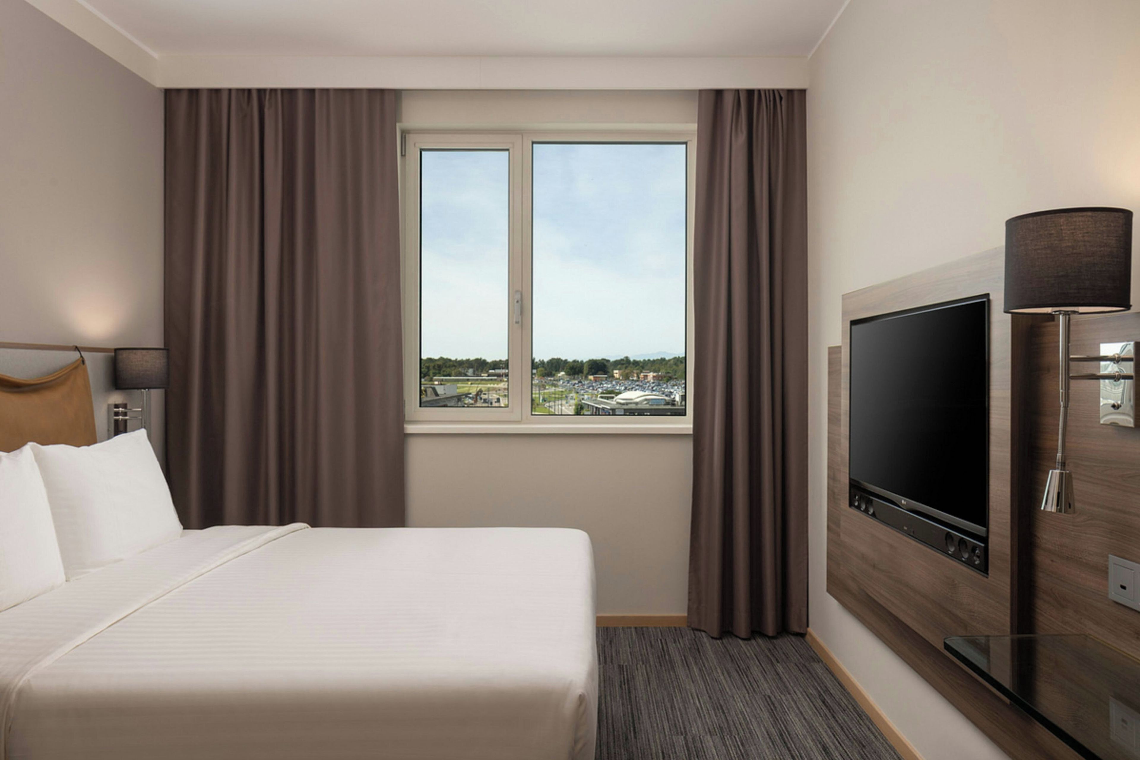 A hotel room with a bed, a television, a window with a view of the city, and curtains.