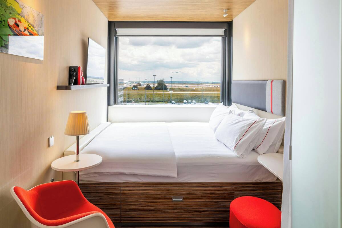 Ein modernes Hotelzimmer mit einem großen Bett, einem kleinen runden Tisch, einem roten Sessel und Blick auf einen Flughafen.