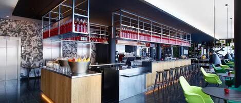 Un moderno bar con una barra alargada, varios estantes llenos de botellas y sillas verdes.
