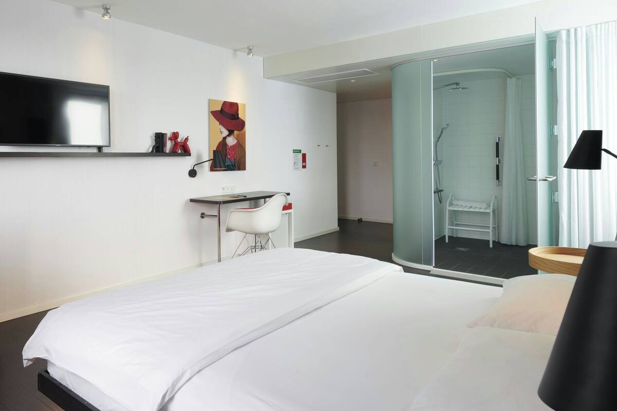 Ein modernes Hotelzimmer mit einem großen Bett, einem Schreibtisch, einem Stuhl, einem Fernseher und einer Dusche.