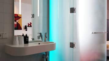 Un baño moderno con puerta de cristal, un lavabo blanco con artículos de aseo y un espejo sobre el lavabo.