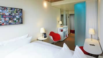 Une chambre d’hôtel avec un grand lit, un fauteuil rouge, une petite table avec une lampe, un téléviseur fixé au mur qui diffuse des affiches de films, et un miroir qui reflète la pièce.