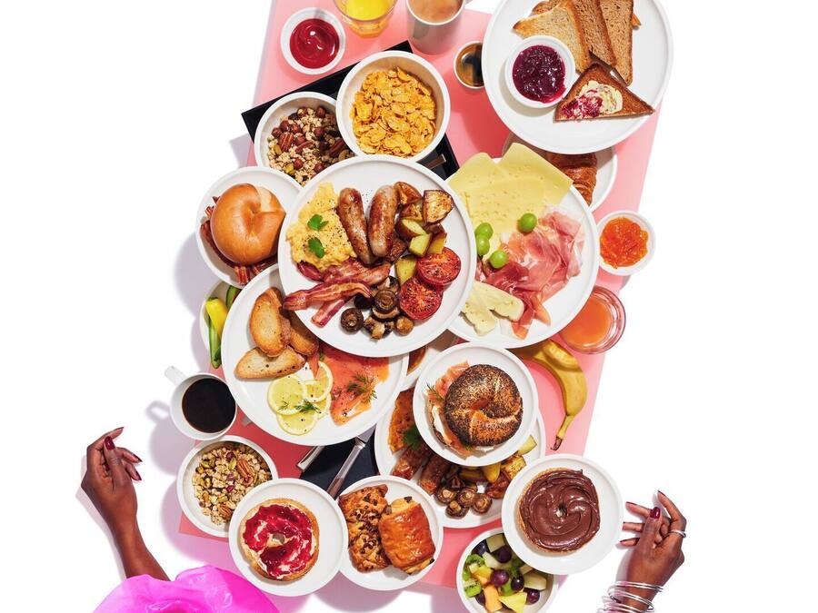 Diversos alimentos de café da manhã dispostos sobre uma mesa, incluindo torradas, ovos, linguiça e frutas.