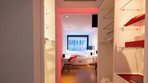 A modern hotel room with a wooden floor, a desk with a chair, a bed, a window, and a closet with hanging clothes.