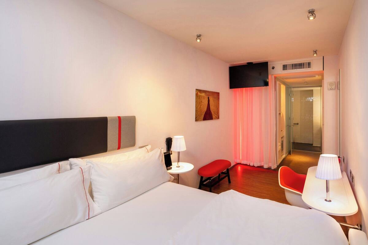 Quarto de hotel com uma cama grande, uma mesa pequena com abajur, uma cadeira vermelha e uma porta que dá para um banheiro.