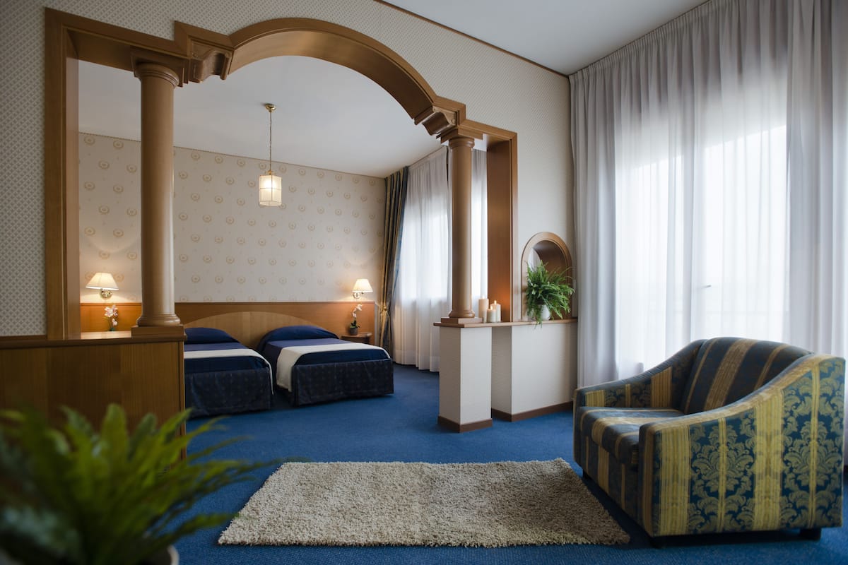 Ein Hotelzimmer mit zwei Betten, einer Sitzecke mit einem Sessel, einem kleinen Tisch und einem großen Fenster mit Vorhängen.