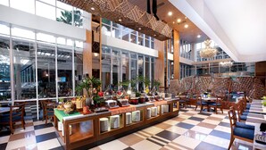 Un amplio vestíbulo de hotel con bufé, piso de tablones de ajedrez, ventanas grandes y un techo decorativo.