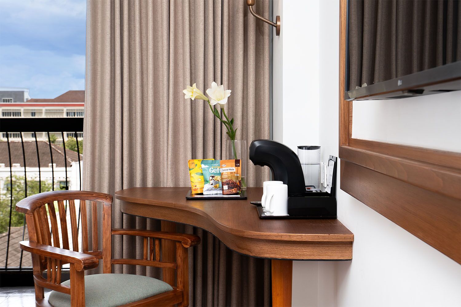 Une chambre d’hôtel avec un bureau en bois, une chaise, une machine à café, un vase avec des fleurs et une vue sur les bâtiments.