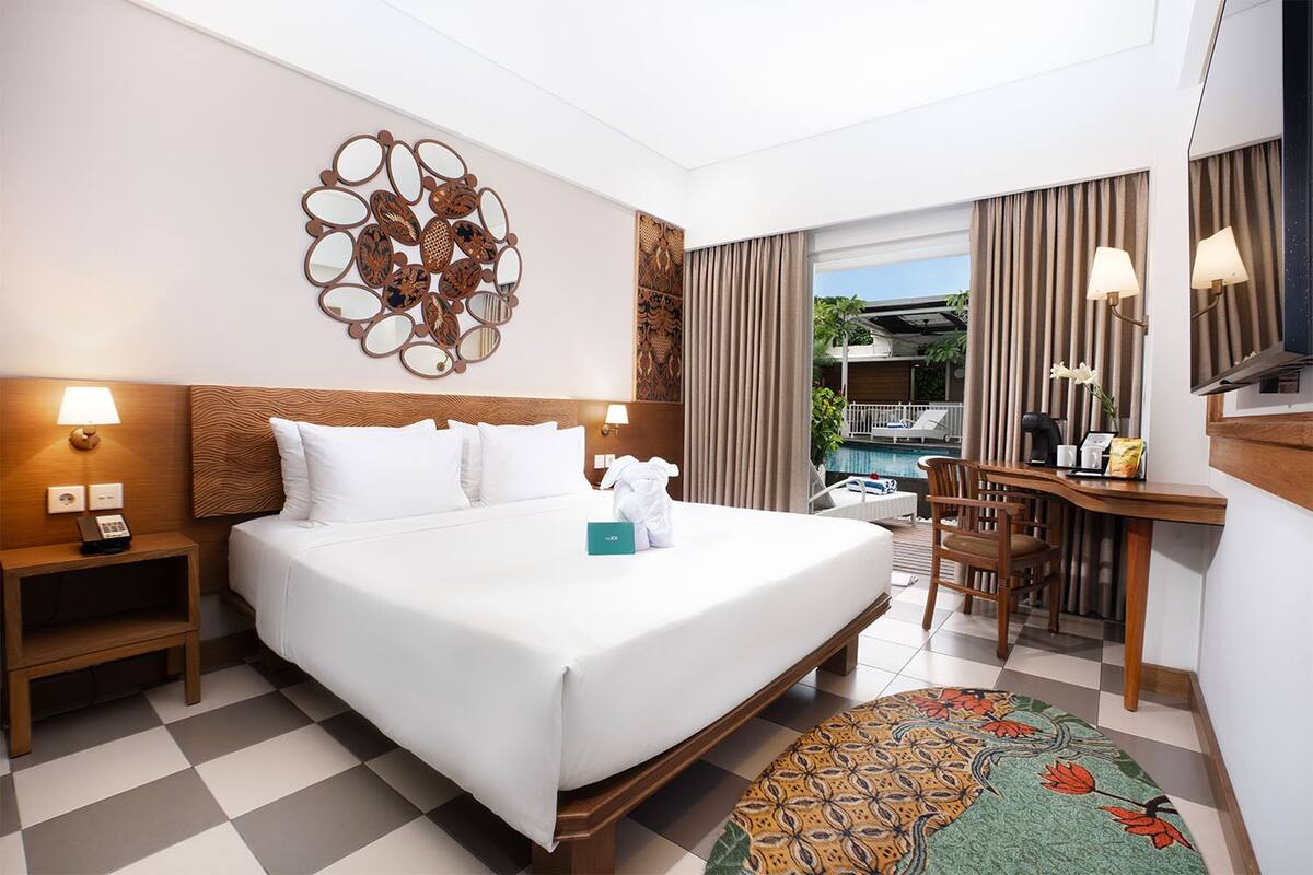 A hotel room with a bed, a desk, a chair, a nightstand, a wall art piece, and a view of an outdoor pool area.