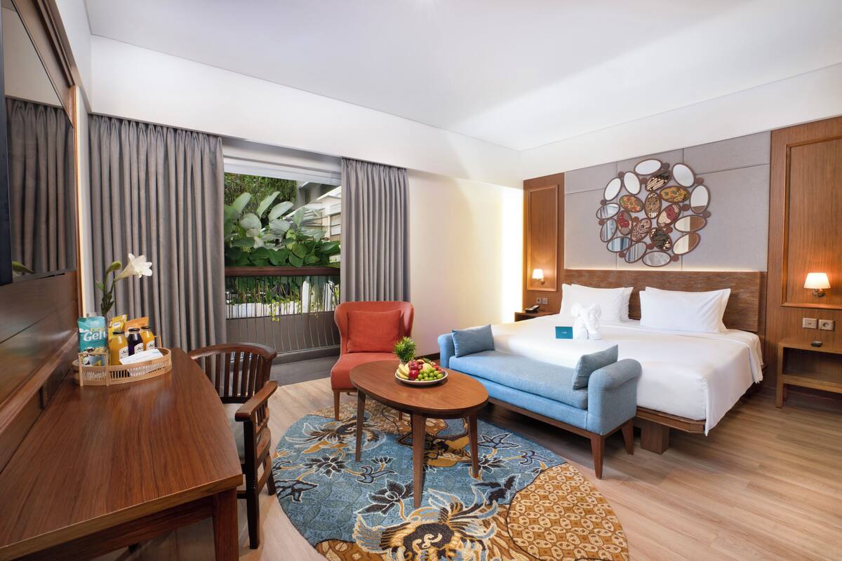 A hotel room with a bed, a sofa, a dining table with a fruit basket, a chair, a small table with a plant, and a view of greenery outside.