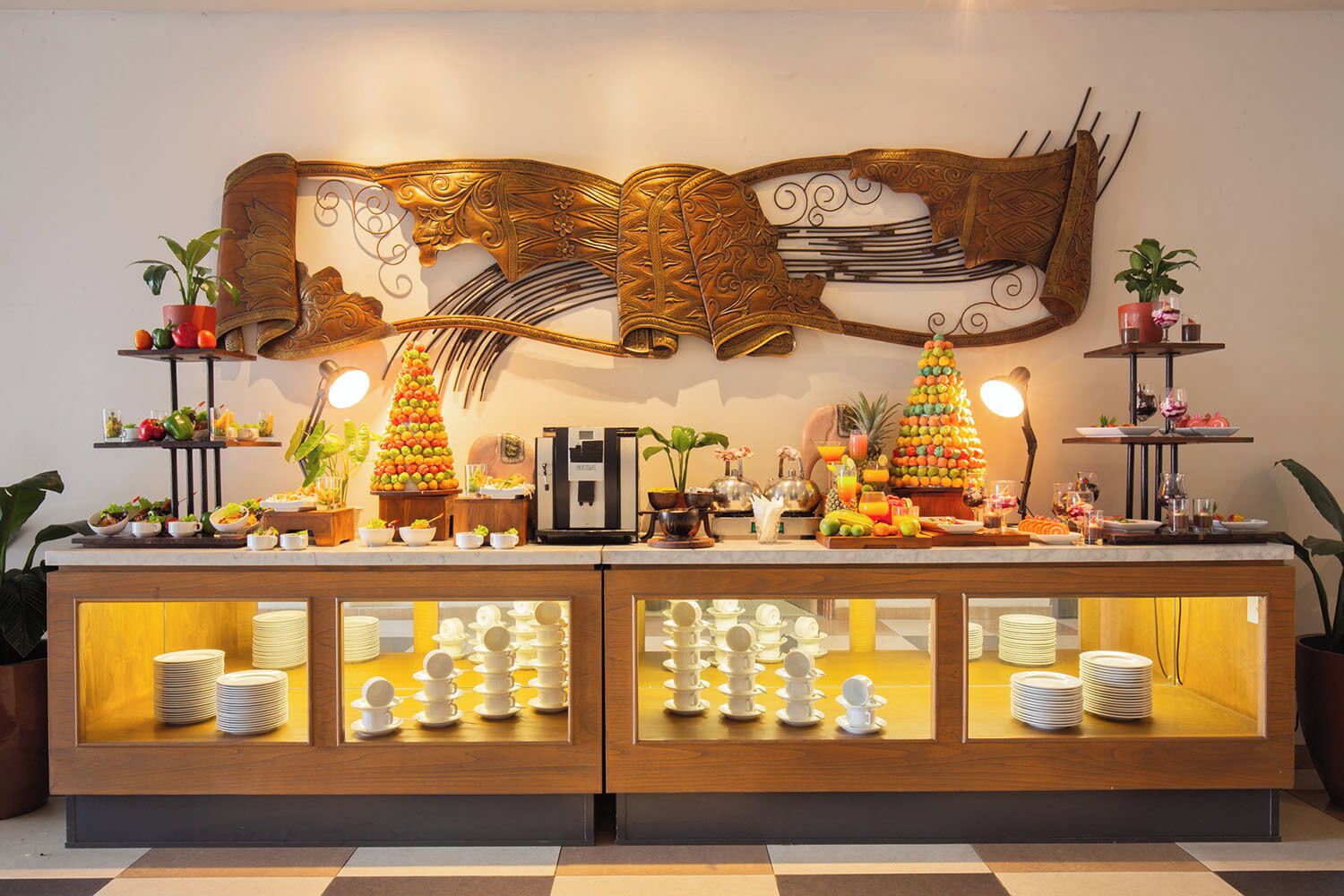 A buffet setup with a decorative wall piece, fruit trees, and a coffee machine.