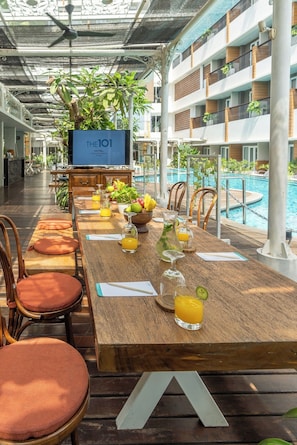 A hotel dining area with a long wooden table set for guests, a pool in the background, and a TV screen displaying 'THE101 HOTEL VILLAVUQUITA'.