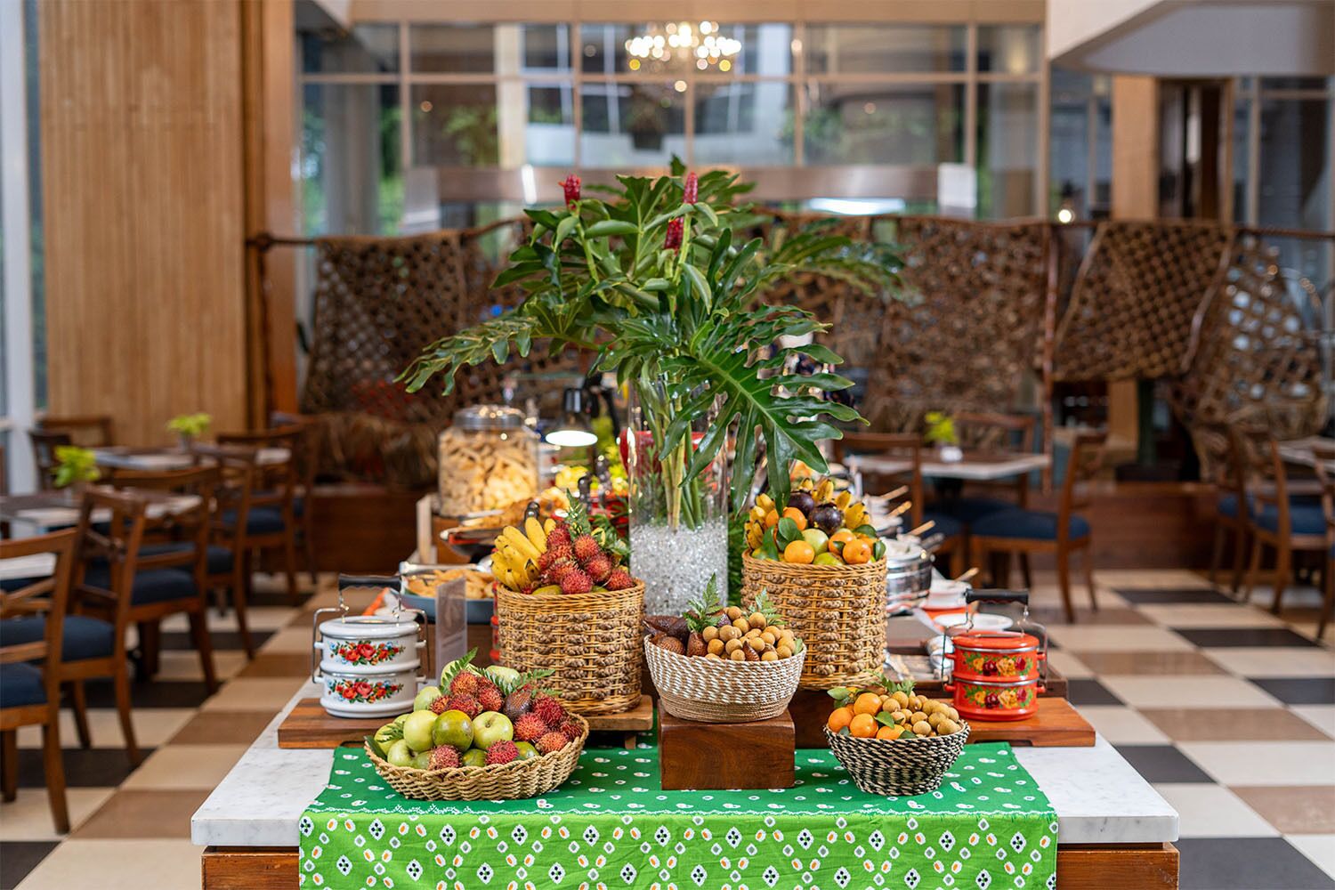 Mesa de buffet con diversas frutas y cestas, una planta en maceta y suelo a cuadros.