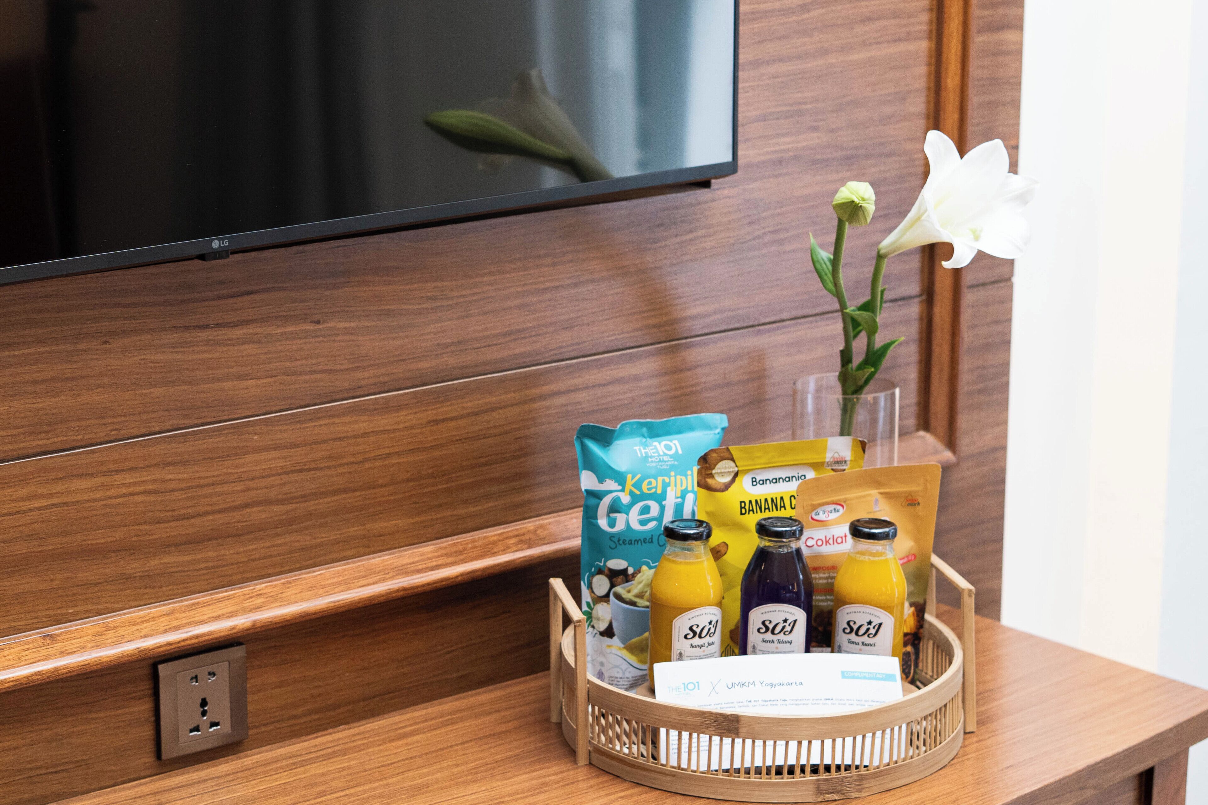 Habitación de hotel con una mesa consola de madera, un televisor de pantalla plana, una bandeja con alimentos para el desayuno y un florero con una flor blanca.
