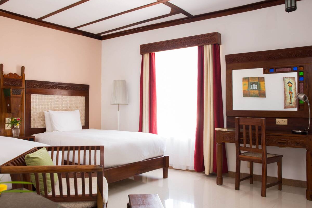 A hotel room with two beds, a desk, a chair, a lamp, and a window with curtains.