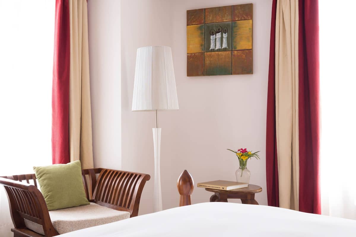 A hotel room with a bed, a wooden chair with a green pillow, a floor lamp, a small table with a book and a vase of flowers, and a painting on the wall.