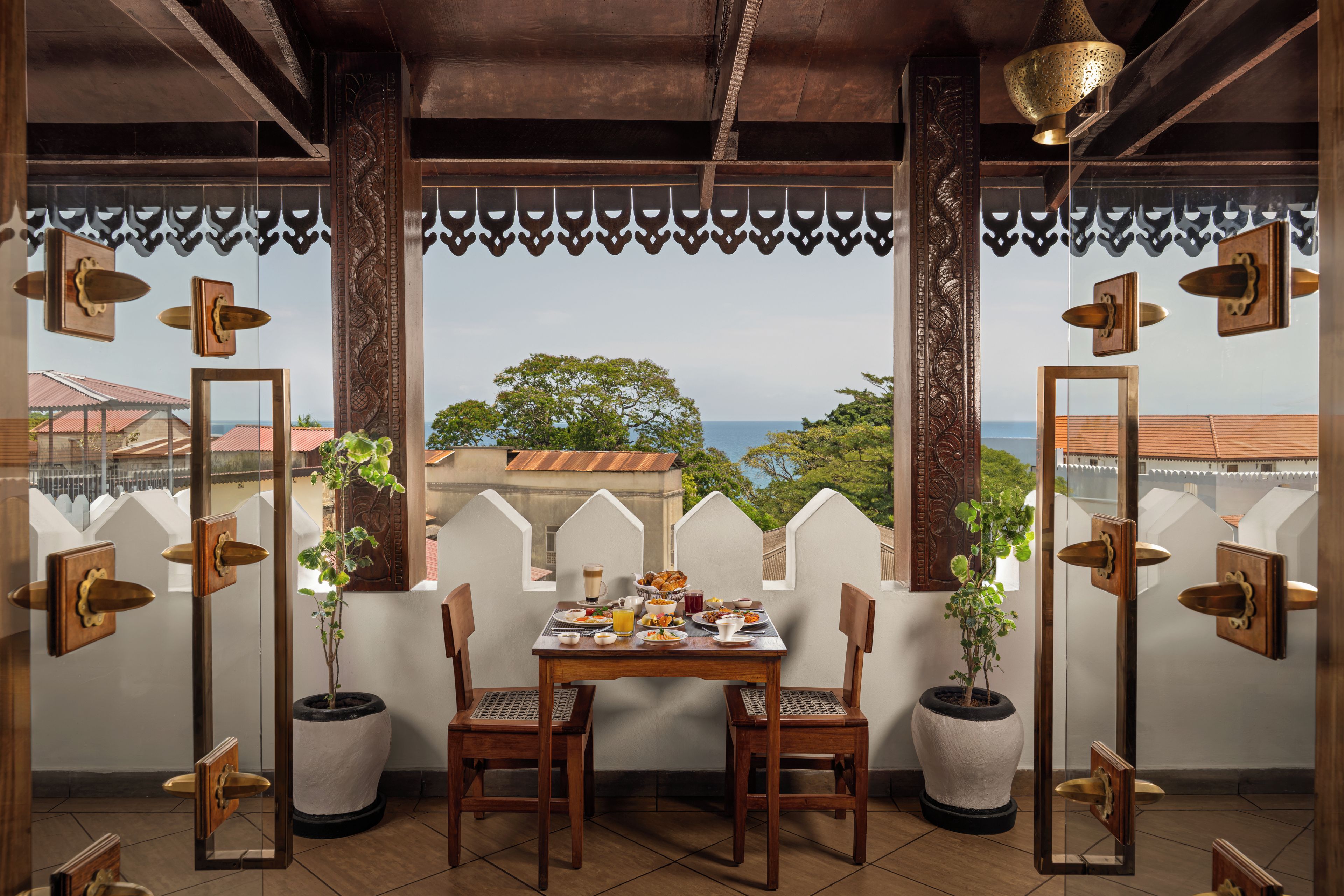 A dining area with a table set for two, chairs, potted plants, and a view of the sea.