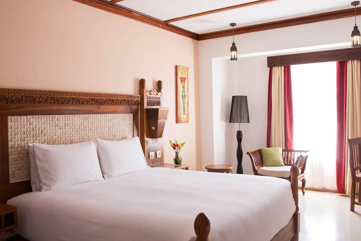 A large bed with a wooden headboard, a bedside table with a vase of flowers, and a chair by the window.