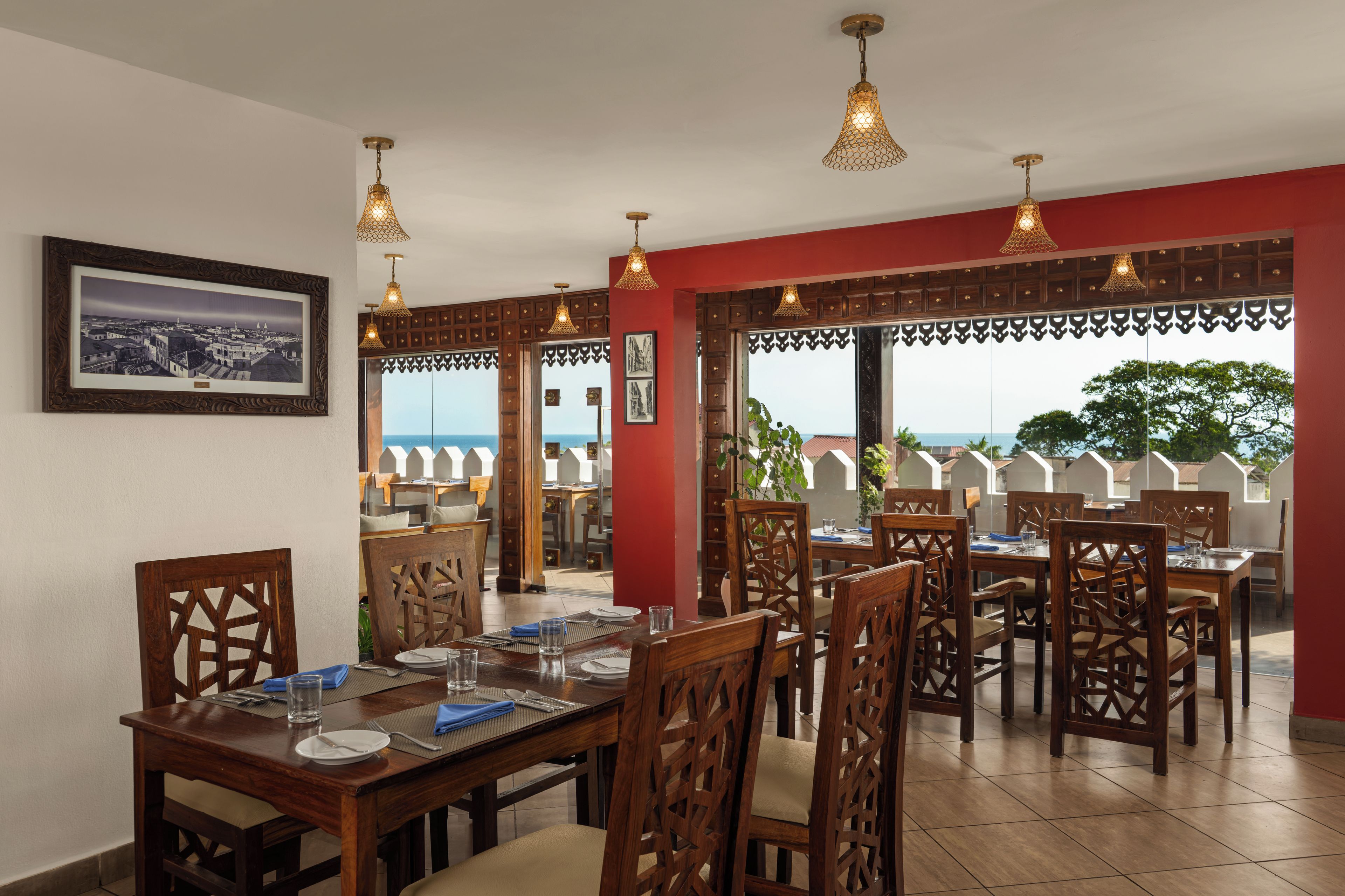 A dining area with wooden furniture and hanging lights, a framed picture on the wall, and a view of the sea through large windows.