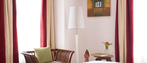 Habitación de hotel con una cama, una silla con cojín, una mesa con un florero y un libro, una lámpara y cortinas.