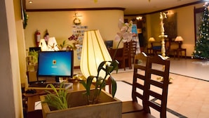 Un vestíbulo de hotel con mostrador de recepción, monitores de computadora, una lámpara grande, una silla de madera y un árbol de Navidad.