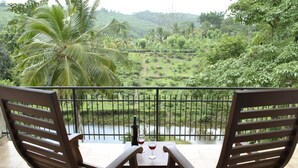 Un balcón con dos sillas de madera y una mesa, con vistas a un paisaje verde y exuberante con un río y palmeras.