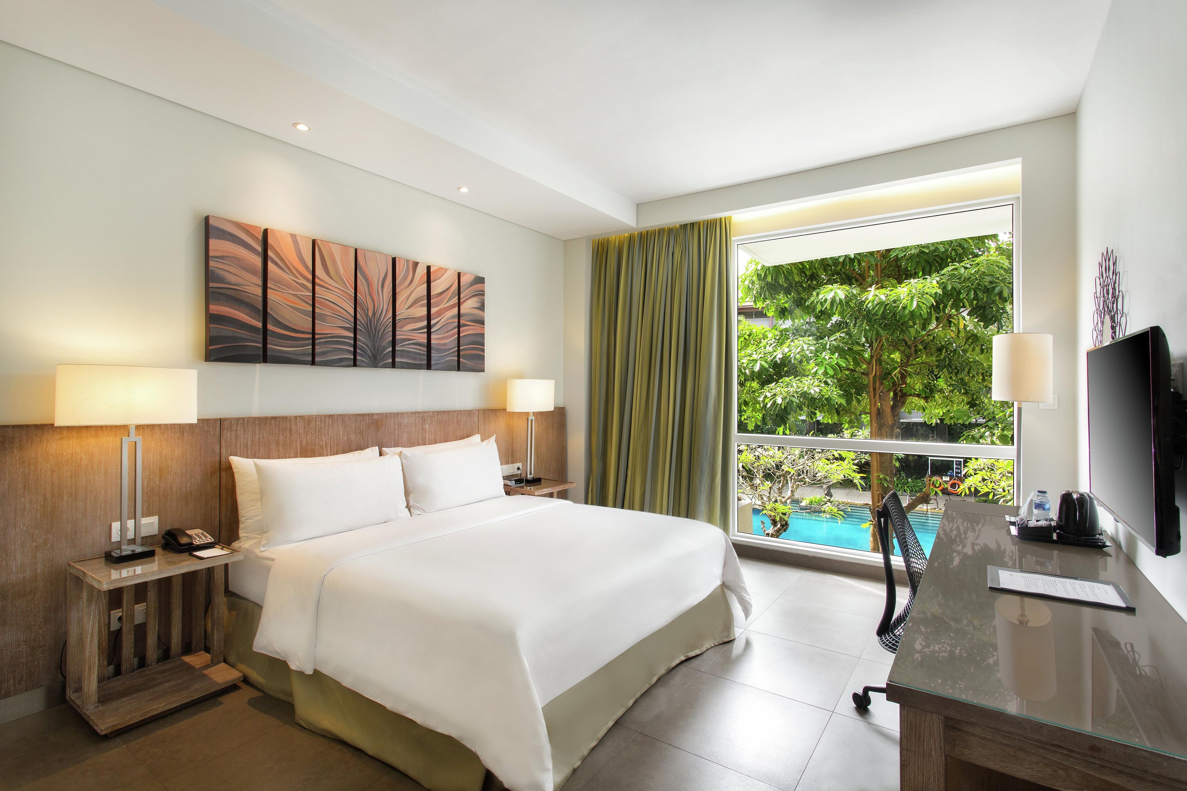 A hotel room with a bed, bedside lamps, a painting, a window with curtains, a desk with a TV, and a view of a swimming pool.