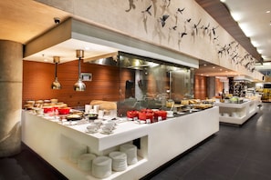 A buffet setup with various food items and utensils, a glass partition, and a wall with a bird mural. - Hilton Garden Inn Bali Ngurah Rai Airport (Tuban)