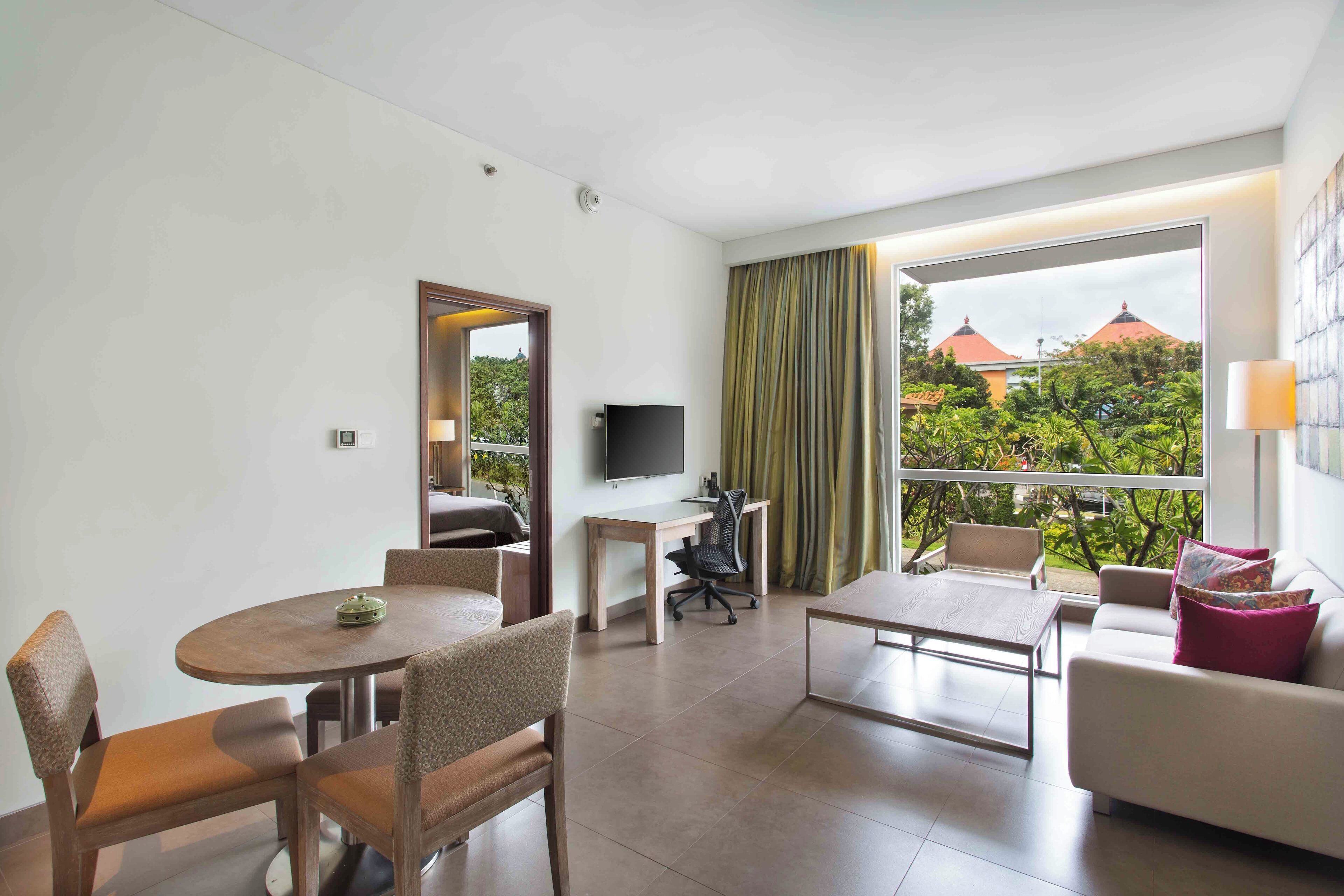 A modern hotel room with a dining area, a sofa, a TV, and a balcony with a view of greenery and buildings.