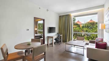 A modern hotel room with a dining area, a sofa, a TV, and a balcony with a view of greenery and buildings.