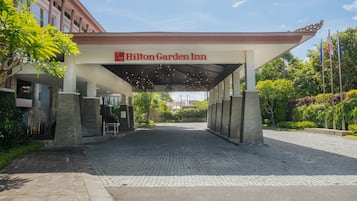The entrance of the Hilton Garden Inn with a covered walkway, paved area, and surrounding greenery.