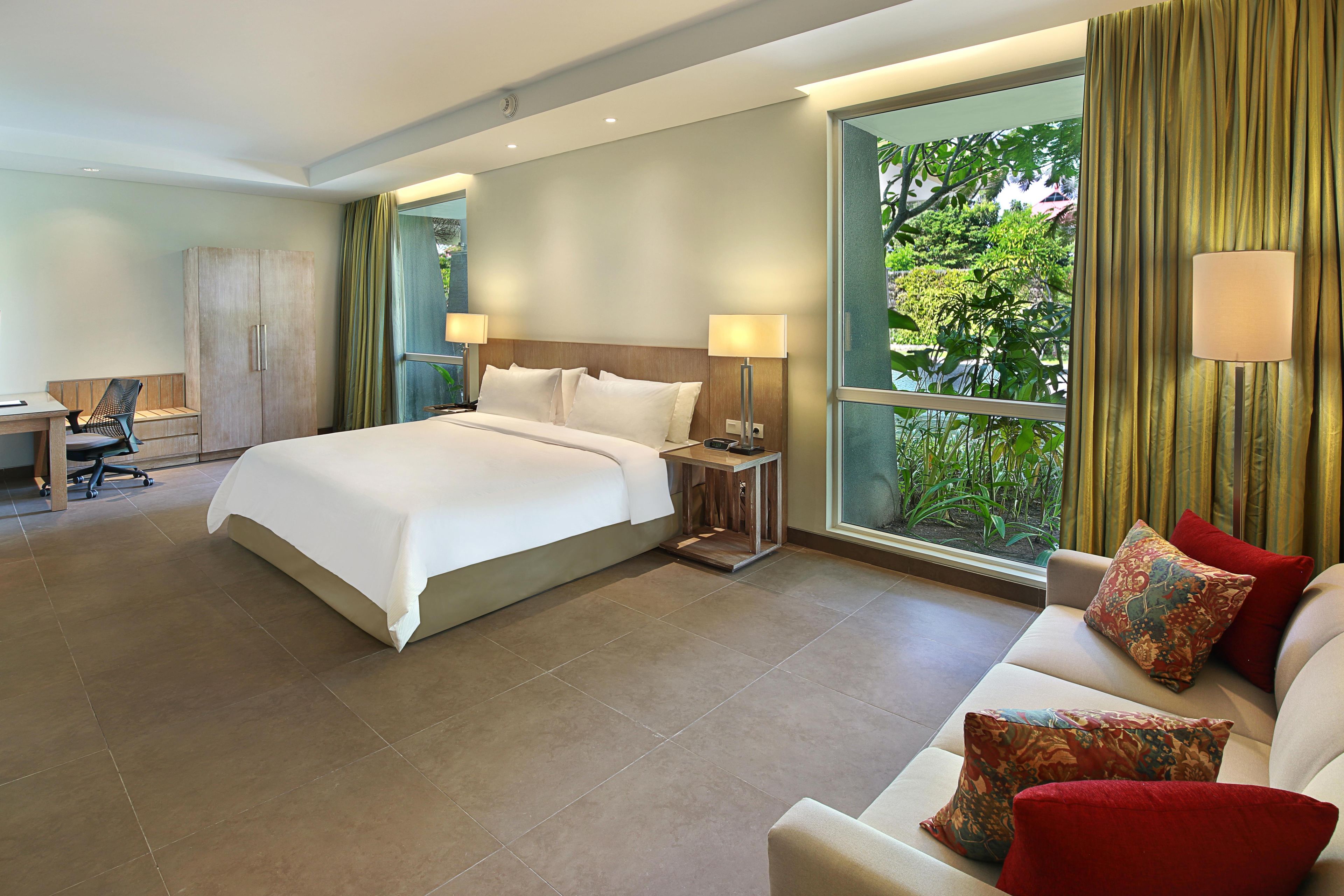 Quarto de hotel com uma cama grande, escrivaninha, poltrona, sofá e janela com cortinas.
