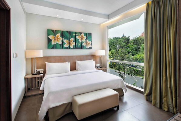 A modern hotel room with a large bed, a bench, a nightstand, a lamp, and a view of greenery through the window. - Hilton Garden Inn Bali Ngurah Rai Airport (Tuban)