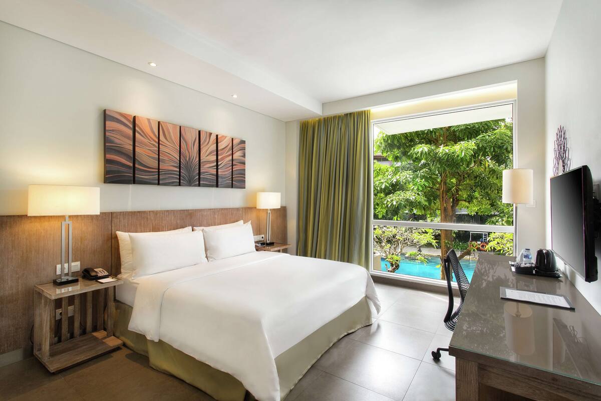 Une chambre d’hôtel équipée d’un lit, d’un bureau, d’une télévision et d’une fenêtre donnant sur une piscine.