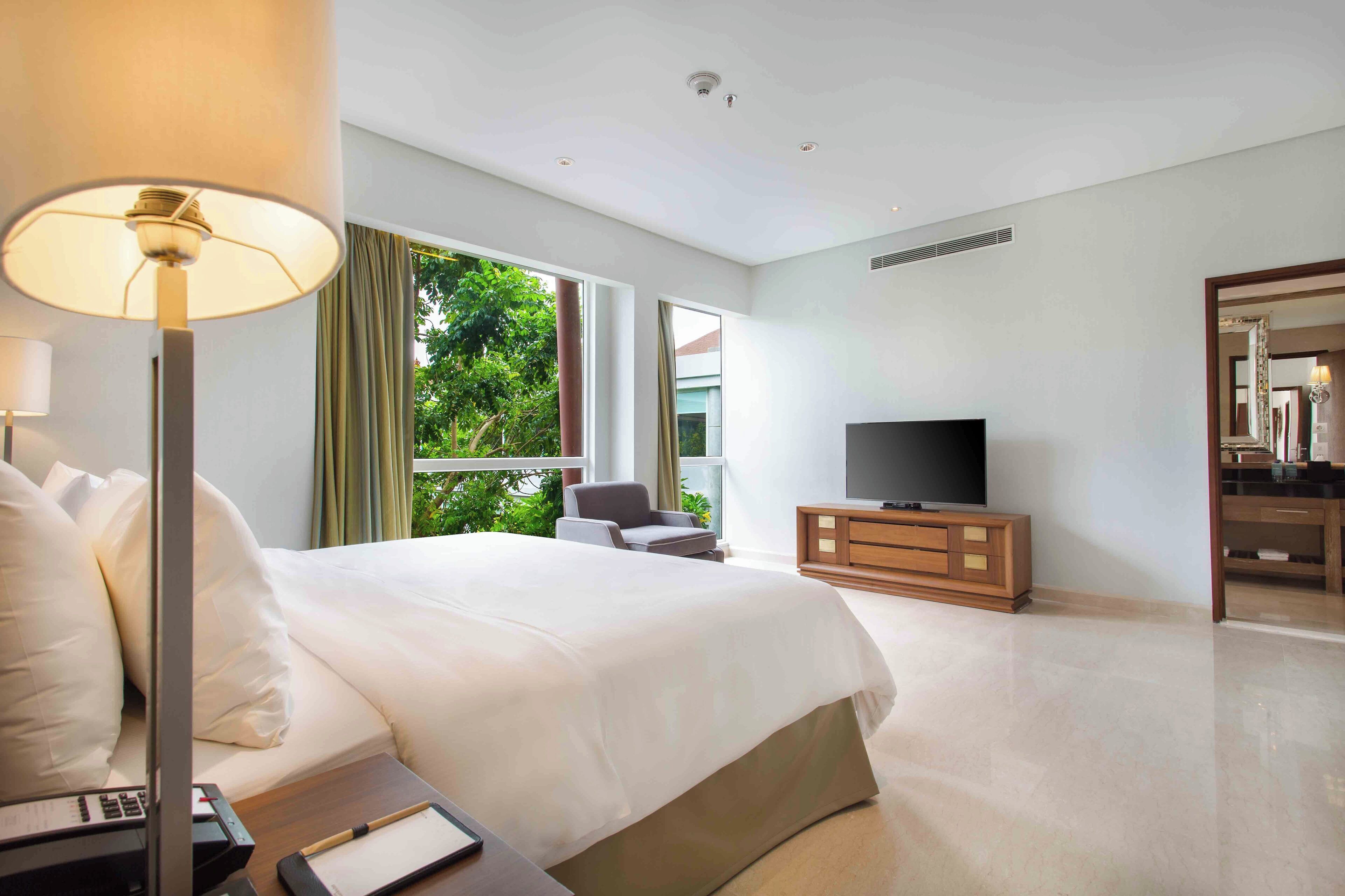 A hotel room with a large bed, a television, a desk with a lamp, and a view of greenery outside.