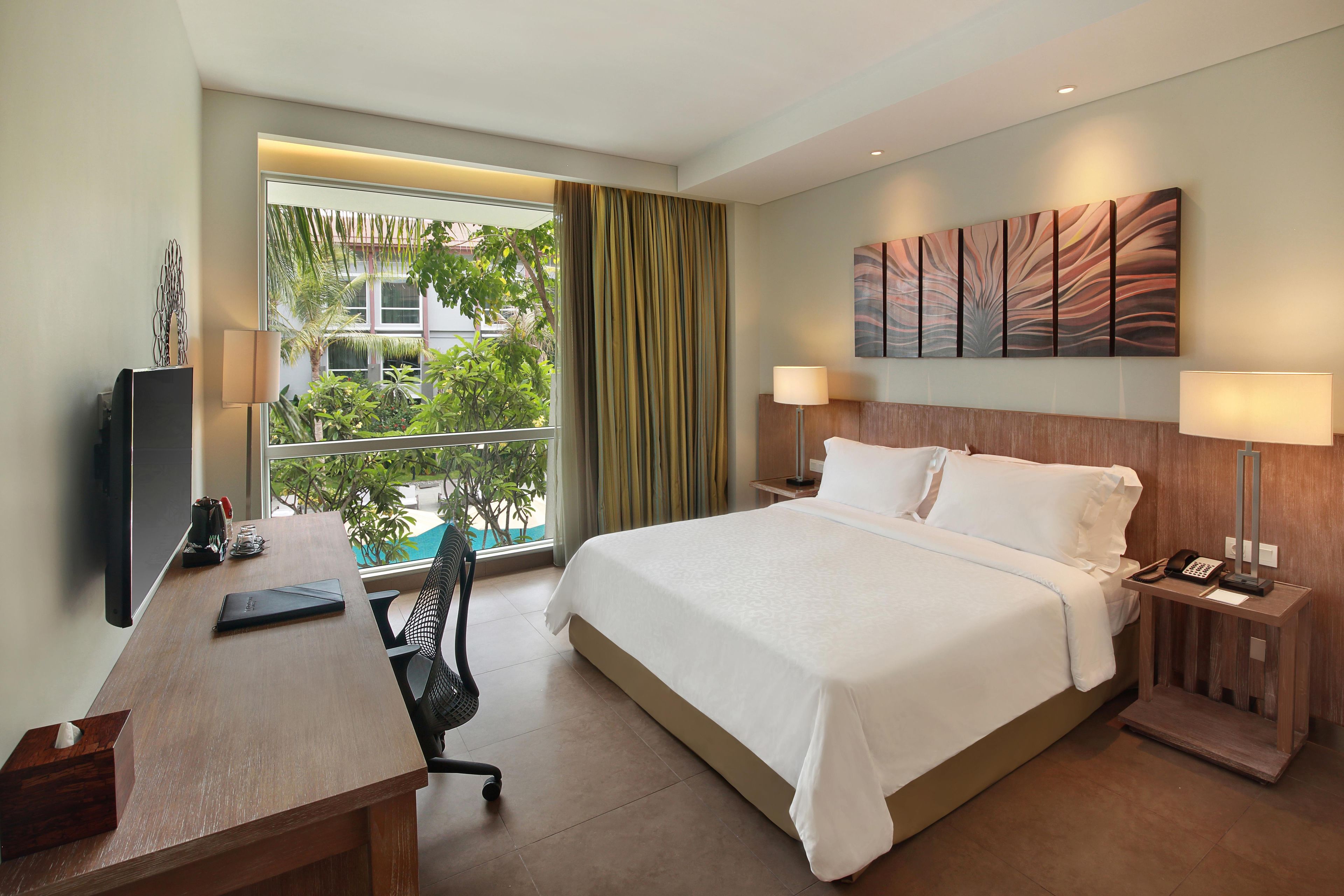 A hotel room with a bed, desk, chair, and a view of greenery outside.