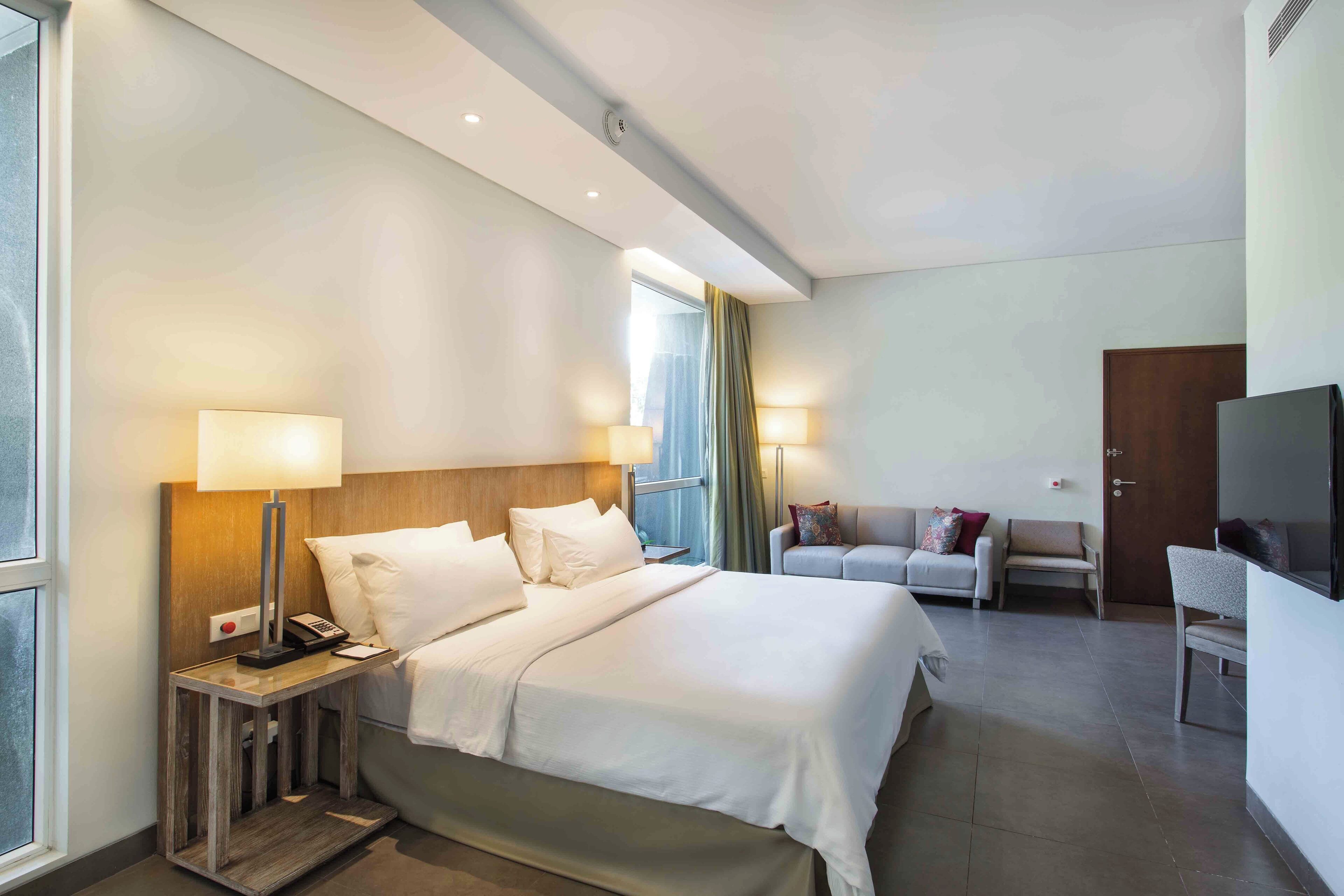 Habitación de hotel con una cama grande, lámparas de noche, un escritorio con teléfono, televisor y un área de descanso con sofá y sillones.