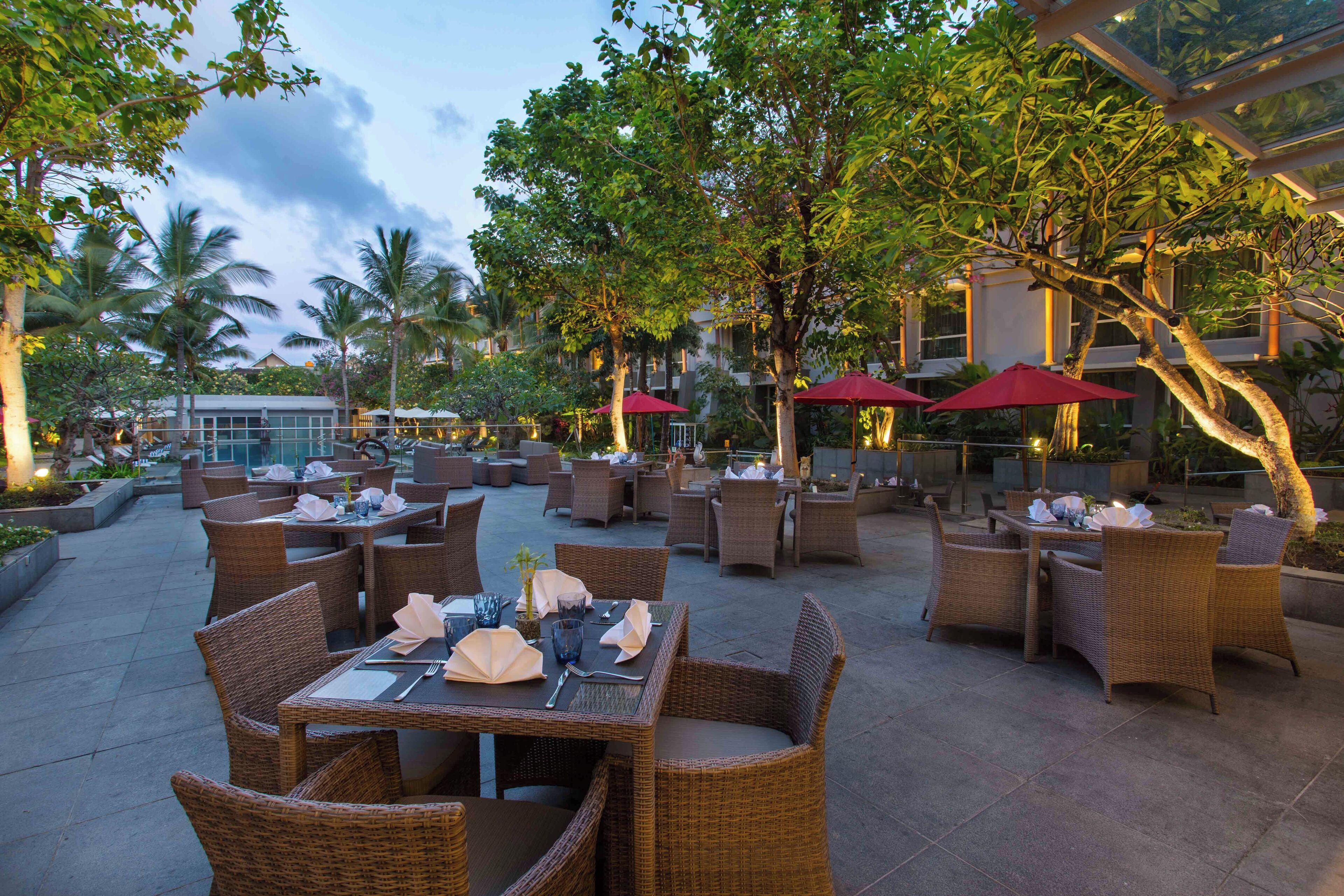 An outdoor dining area with wicker furniture and red umbrellas, surrounded by trees and buildings.