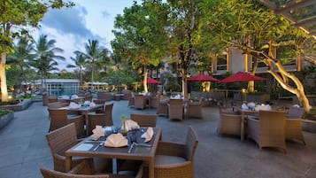 An outdoor dining area with wicker furniture and red umbrellas, surrounded by trees and buildings.