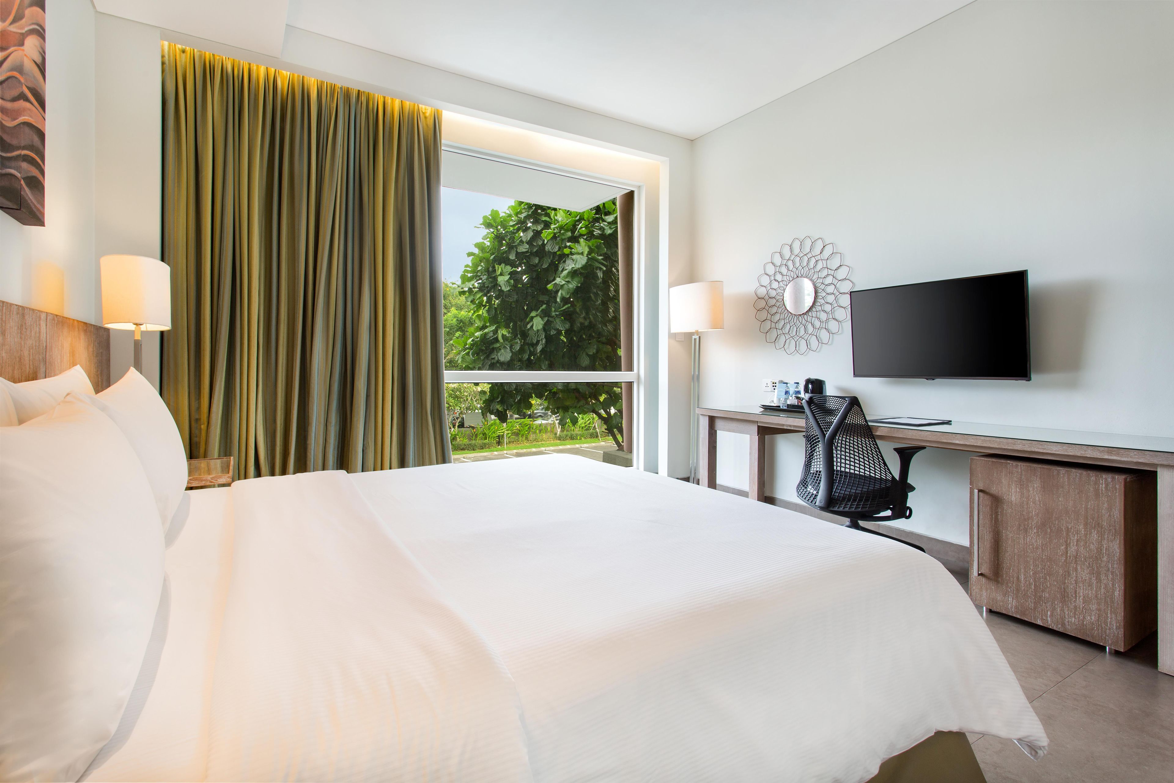 A hotel room with a bed, a desk with a chair, a television, and a window with curtains.