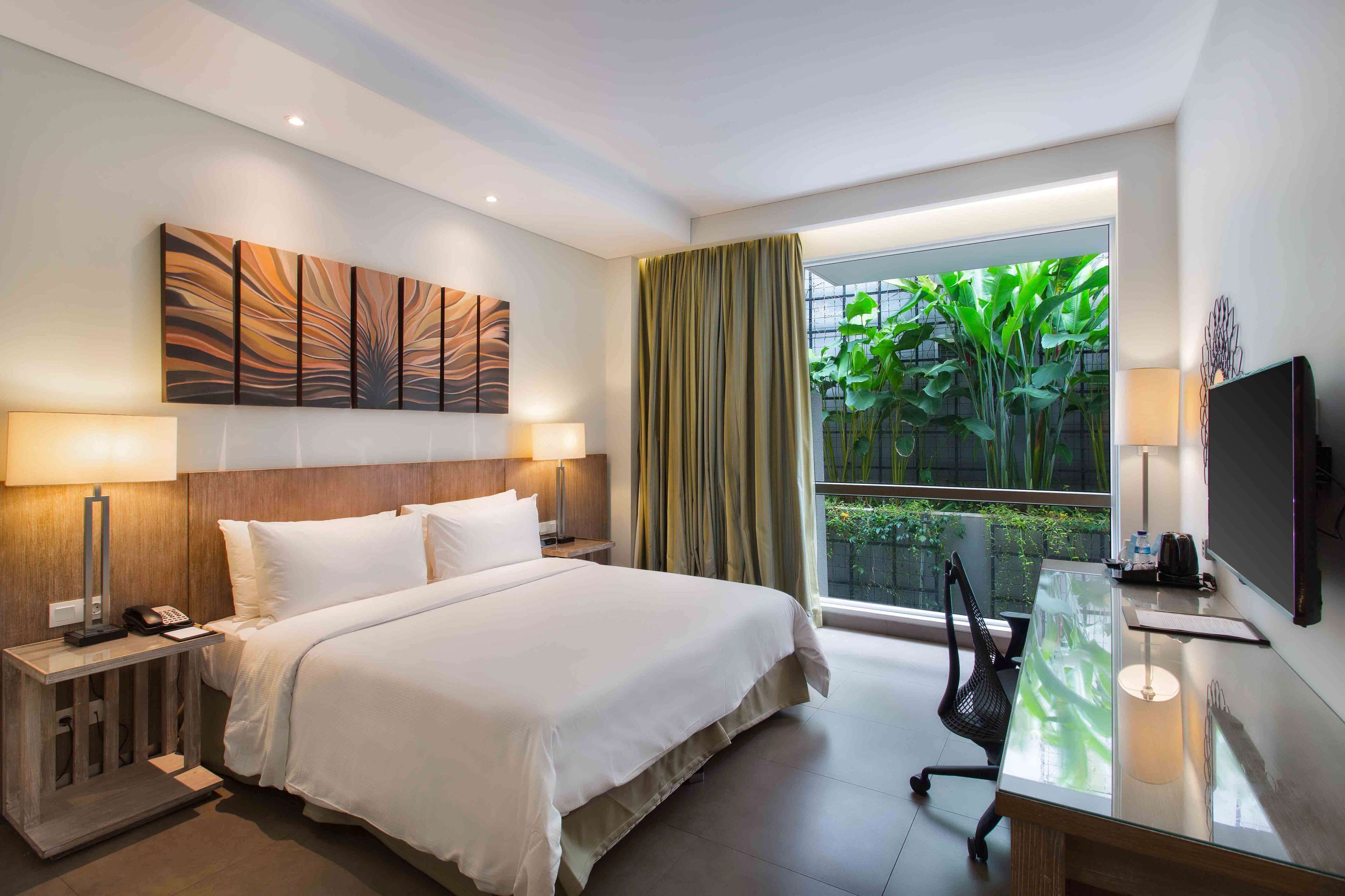 A hotel room with a large bed, bedside lamps, a desk with a chair, a TV, and a window with curtains.