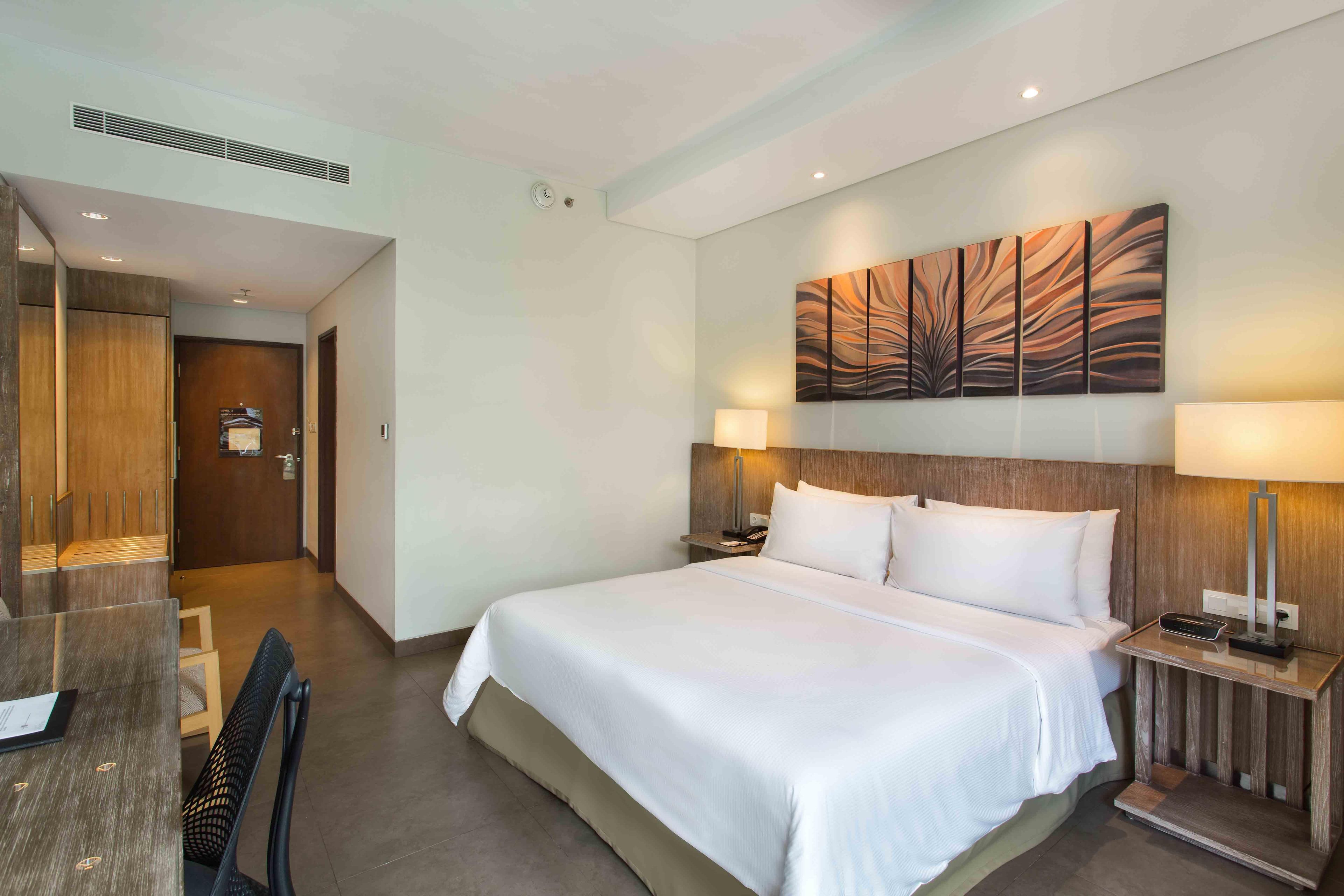 A hotel room with a large bed, two bedside tables with lamps, a headboard, a wall-mounted artwork, and a door leading to a hallway.