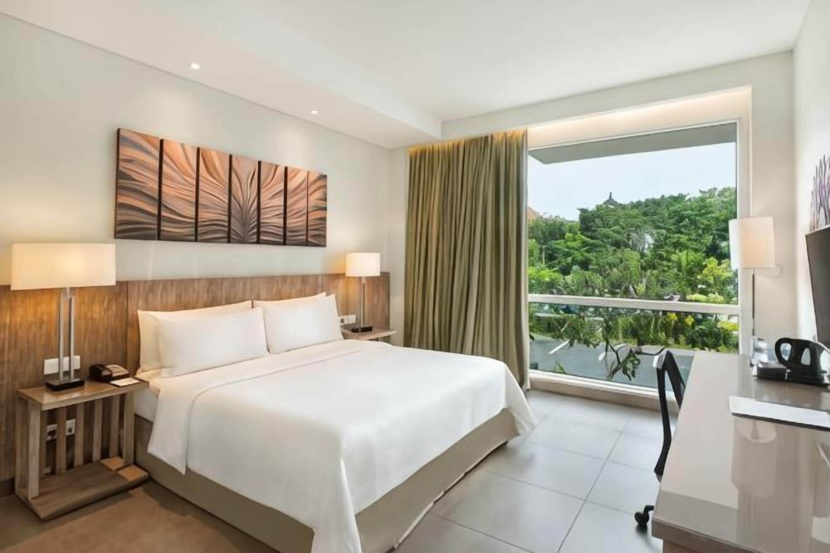 A hotel room with a large bed, two bedside tables with lamps, a desk with a chair, and a view of greenery through a window.
