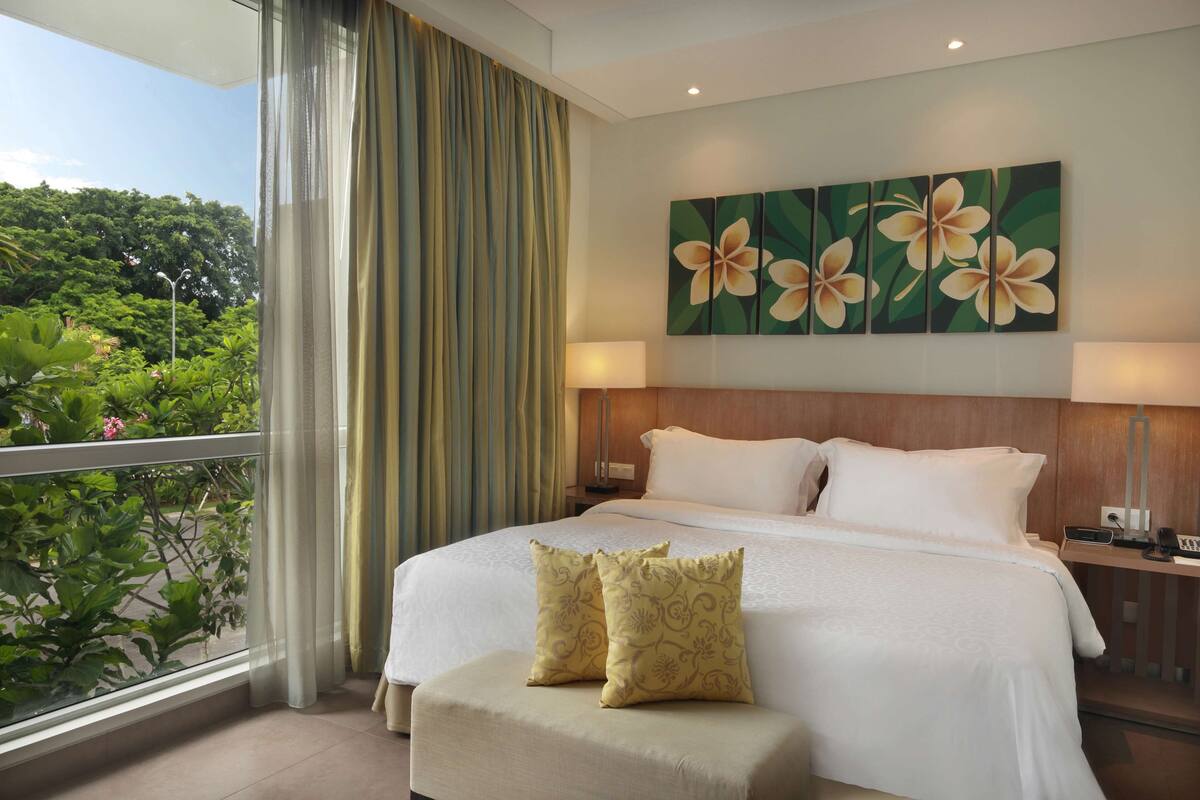 Une chambre d’hôtel avec un grand lit, une vue sur la verdure, un tableau de fleurs accroché au mur et un balcon avec des rideaux.