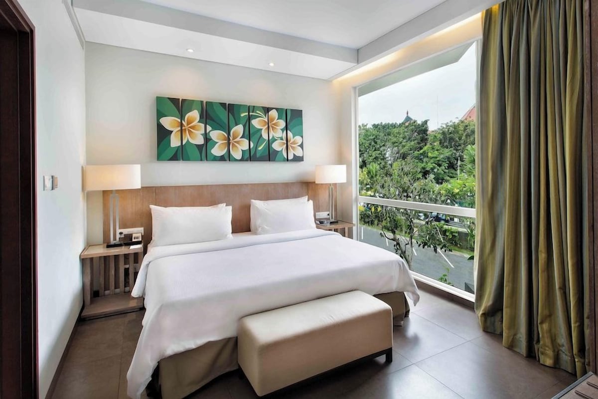 Habitación de hotel con una cama grande, dos mesitas de noche, un banco y vistas a zonas verdes a través de un balcón, adem ás de un cuadro con motivos florales en la pared.