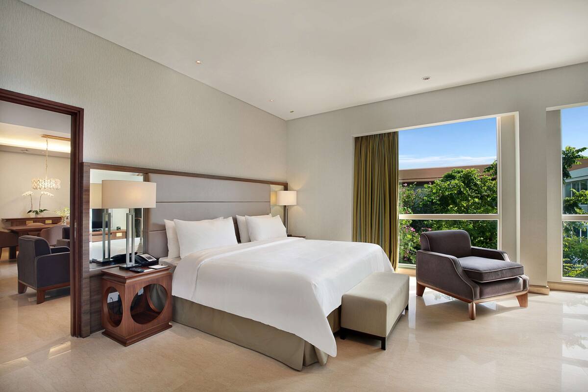 Une chambre d’hôtel moderne, dotée d’un grand lit, d’un bureau avec une lampe, d’une chaise et offrant une vue sur la verdure par la fenêtre.