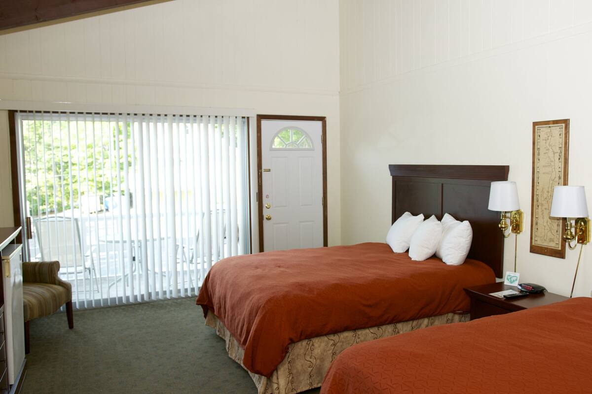 A hotel room with two beds, a desk, a chair, and a balcony with blinds.