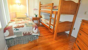 Una camera da letto con un letto a castello in legno, un letto singolo con copriletto, un comodino con lampada e una cassettiera.