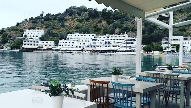 Um restaurante à beira-mar com mesas ao ar livre, vista para a água, edifícios brancos ao fundo e uma paisagem montanhosa.