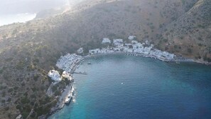 An aerial view of a coastal town with white buildings arranged along the shoreline, a pier extending into the water, and a mountainous backdrop. - Kyma (Sfakia)